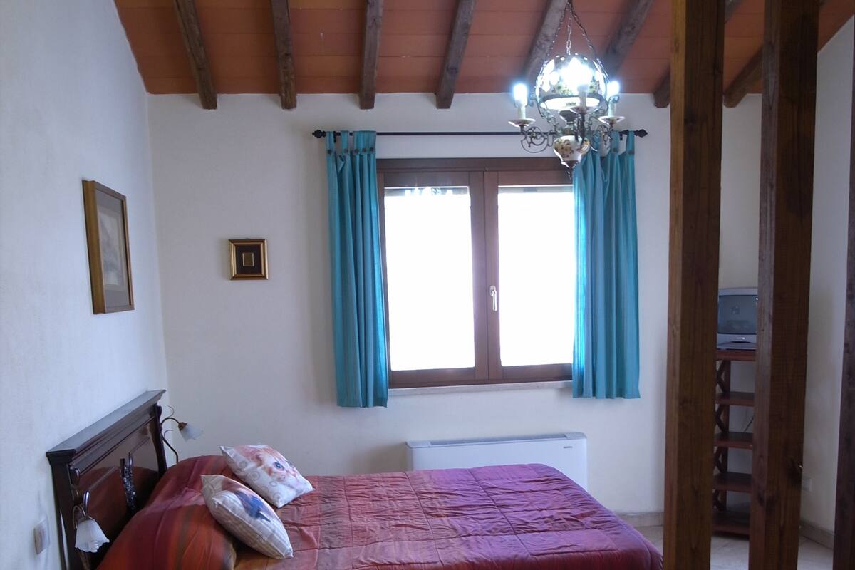 Een slaapkamer met een houten plafond, een bed met een rode sprei, een nachtkastje, een raam met gordijnen en een kroonluchter.