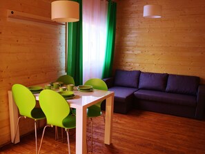 A wooden interior with a dining table, green chairs, a purple sofa, and two pendant lights.