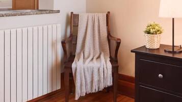 Una habitación de esquina con piso de madera, un radiador, una silla con una manta, una mesita con una lámpara y una planta, y una ventana con persianas.