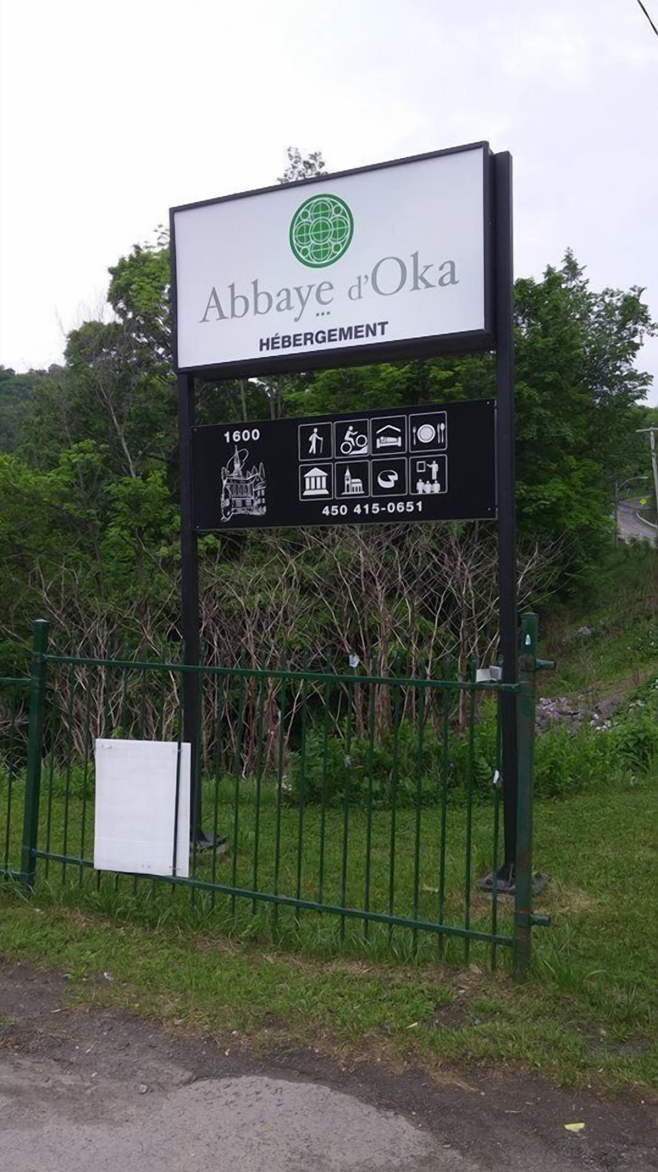 Skilt for Abbaye d'Oka med kontaktnummer og symboler der angiver faciliteter som restaurant, parkering og adgang for handicappede.