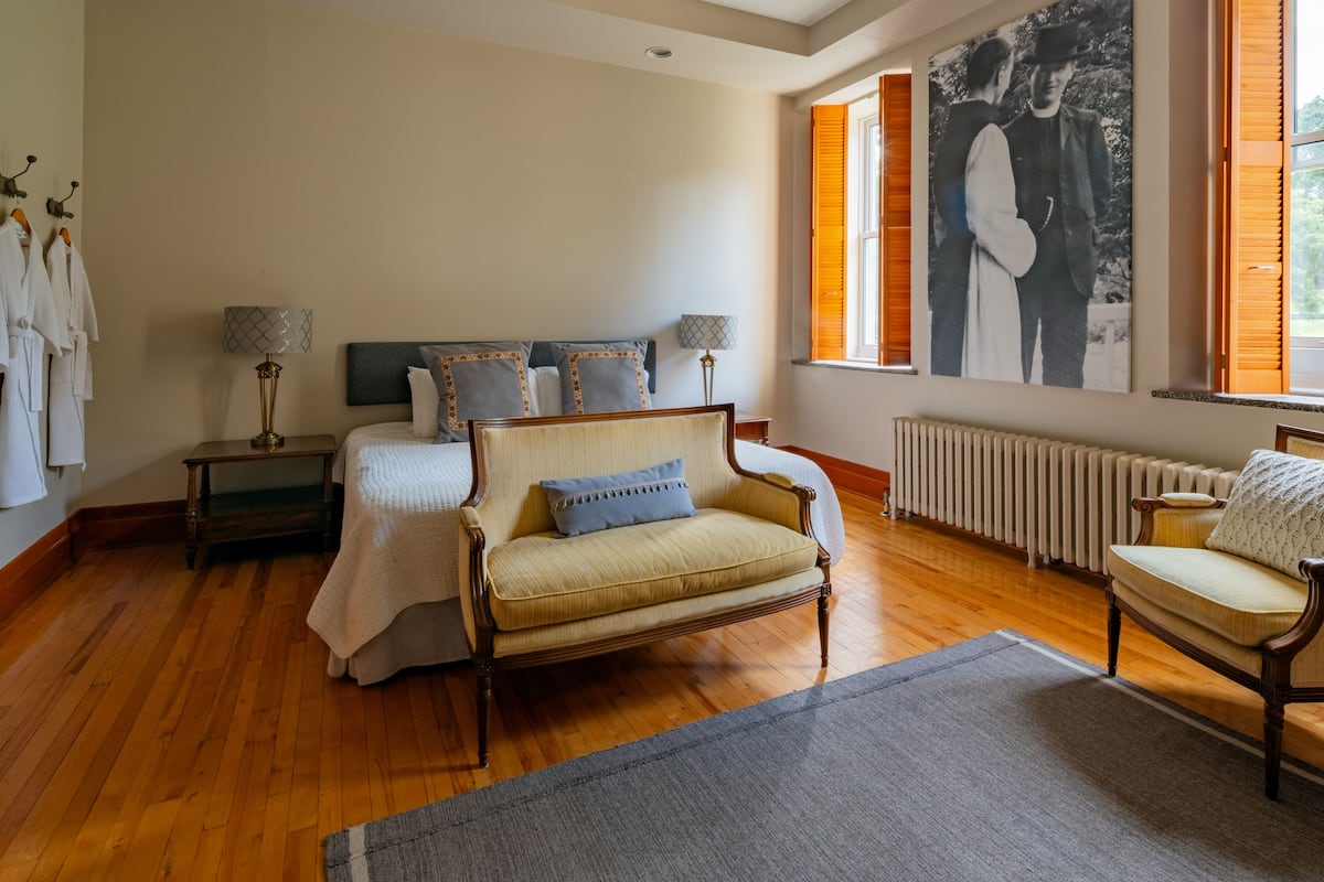 Ein Schlafzimmer mit einem Bett, einer Couch, einem Sessel, einem Heizkörper und einem großen Fenster mit Rollläden.