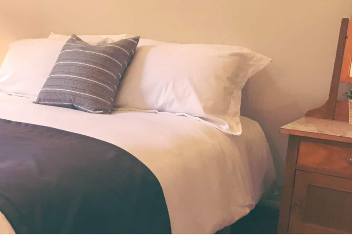 Ein ordentlich bezogenes Bett mit einer dunkelgrauen Tagesdecke, weißen Kissen und einem gestreiften Kissen. Ein Nachttisch mit Lampe und einer kleinen Pflanze ist vorhanden.