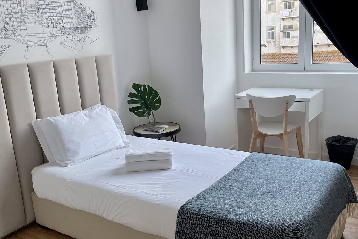 A neatly made bed with a white headboard, a bedside table with a plant, a desk with a chair, and a window with a view of buildings.