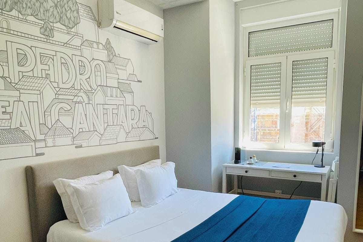 A neatly made bed with white and blue bedding, a grey headboard, and a wall mural depicting a cityscape.