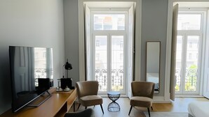 A modern living room with a flat-screen TV, a wooden desk, two armchairs, a small round table, and a large window with a view of buildings. - Lisbon Check-In Guesthouse (Lisbon)