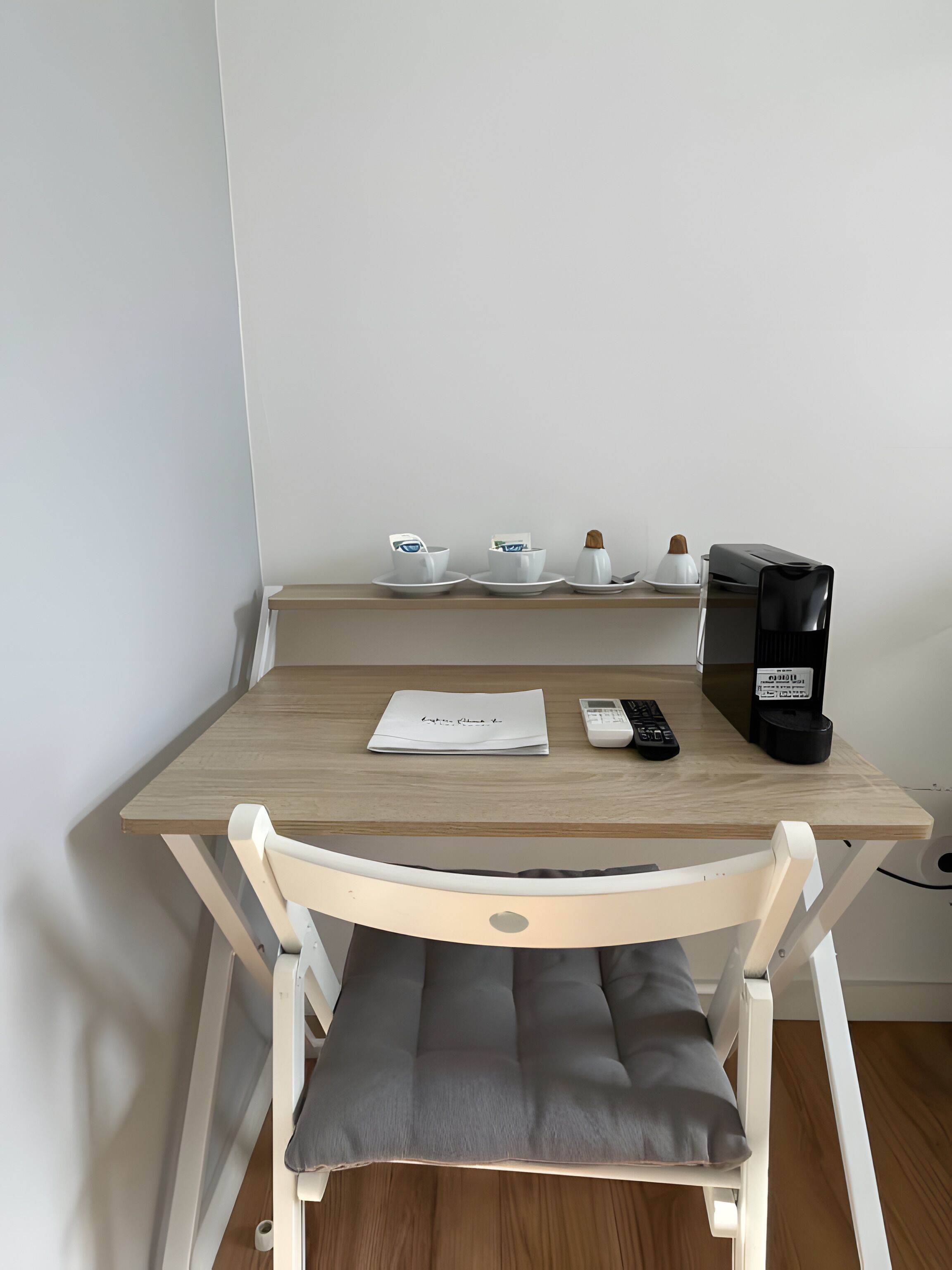 A wooden desk with a white chair, a coffee maker, a remote control, and a piece of paper with a name on it.
