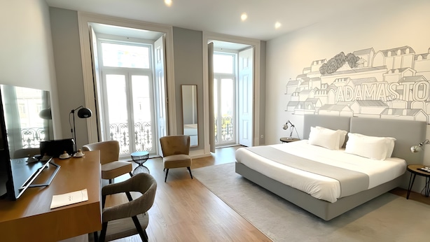 A modern hotel room with a large bed, a desk, two chairs, and a cityscape mural on the wall. - Lisbon Check-In Guesthouse (Lisbon)