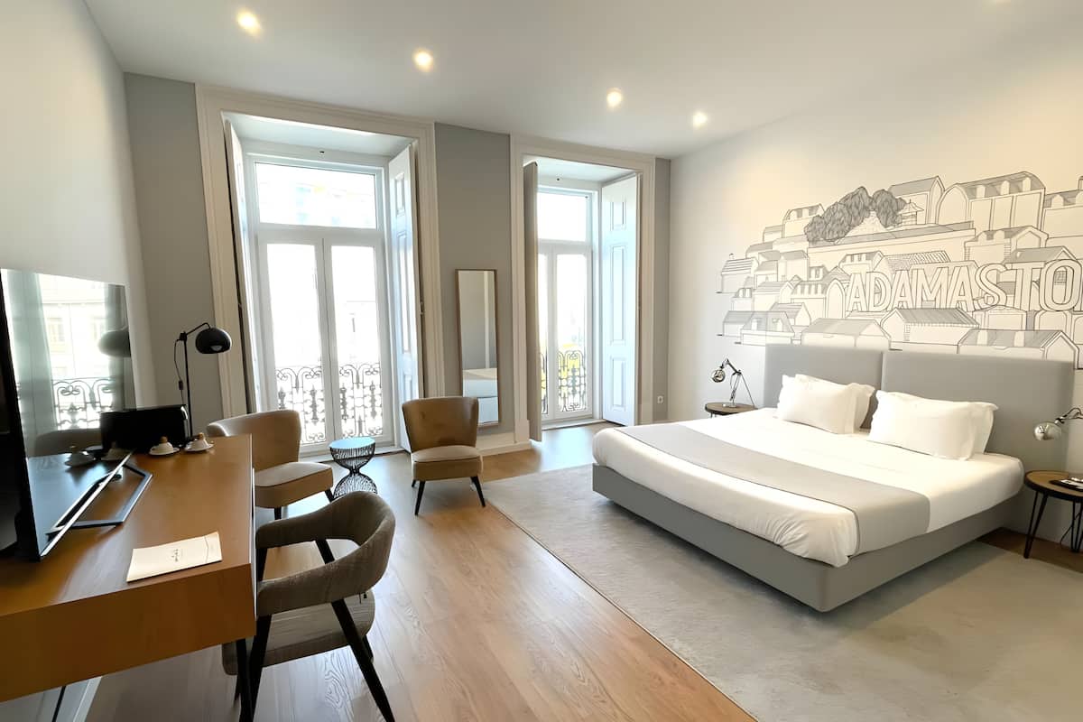 A modern hotel room with a large bed, a desk, two chairs, and a cityscape mural on the wall.