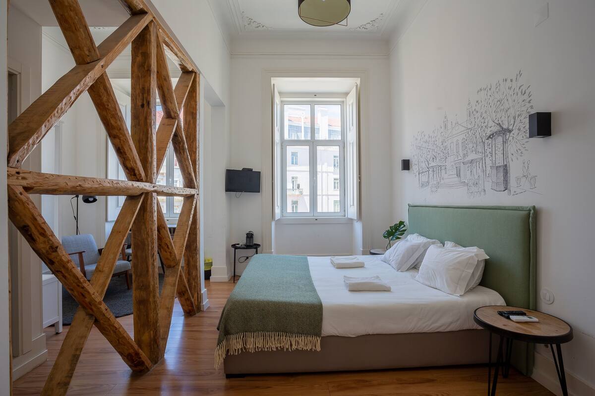 A bedroom with a bed, a green headboard, a window, and a wall with a sketched cityscape.
