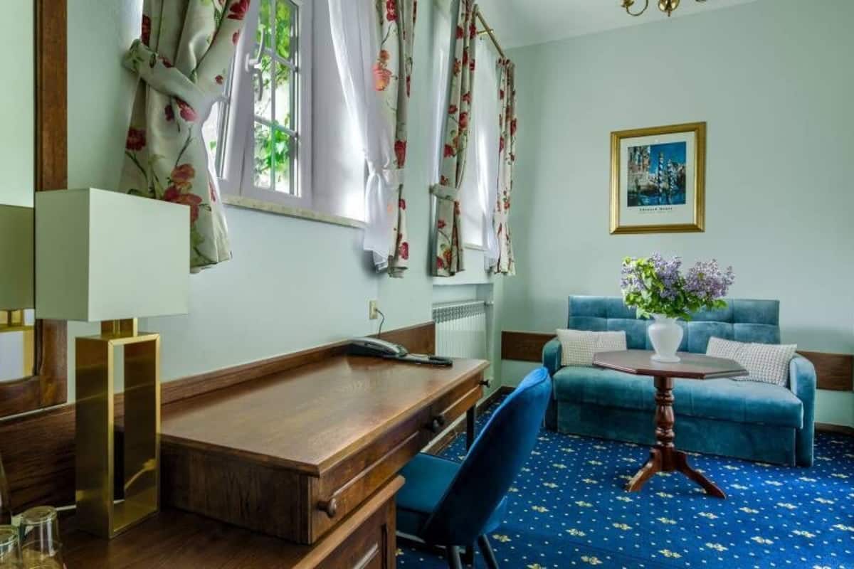 Quarto com escrivaninha de madeira, sofá azul, mesa redonda pequena com um vaso e um espelho grande.
