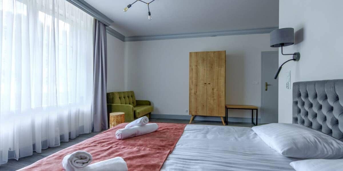 A hotel room with a bed, a green armchair, a wooden wardrobe, and a small table.