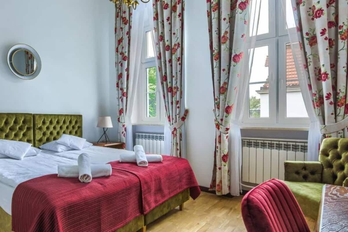 Quarto com cama estofada verde, lençóis brancos, manta vermelha, mesa de cabeceira com abajur e cortinas florais.