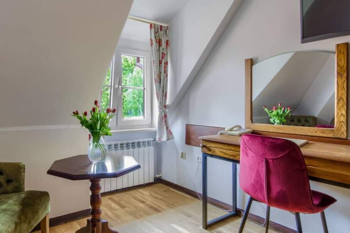 Um quarto com um sofá verde estofado, uma pequena mesa de madeira com um vaso de flores, uma cadeira vermelha, uma escrivaninha de madeira com um espelho e uma janela com cortinas.