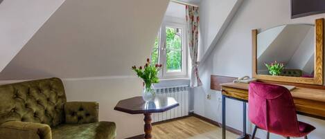 Une pièce avec un canapé vert à motifs, une petite table en bois sur laquelle est posé un vase de fleurs, une chaise rouge, un bureau en bois avec un miroir et une fenêtre avec des rideaux.