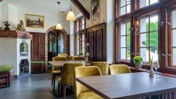 A dining area with a wooden table and yellow chairs, large windows with a view of greenery, and a brick fireplace.