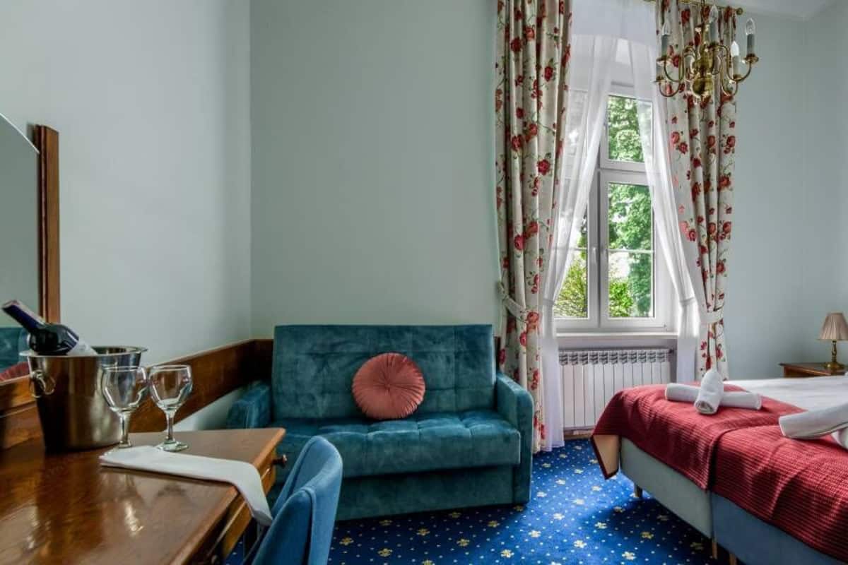 Quarto com cama, sofá azul, mesa de madeira com copos e janela com cortinas florais.