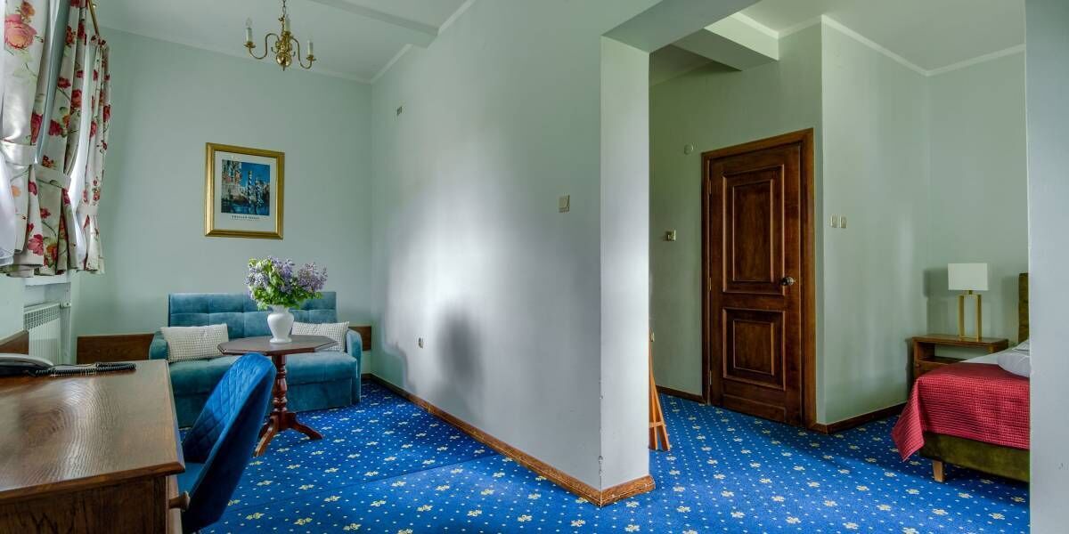 Ein Zimmer mit blauem, gemustertem Teppich, einem blauen Sofa, einem Holztisch, einer Holztür und einem Bett mit roter Decke.