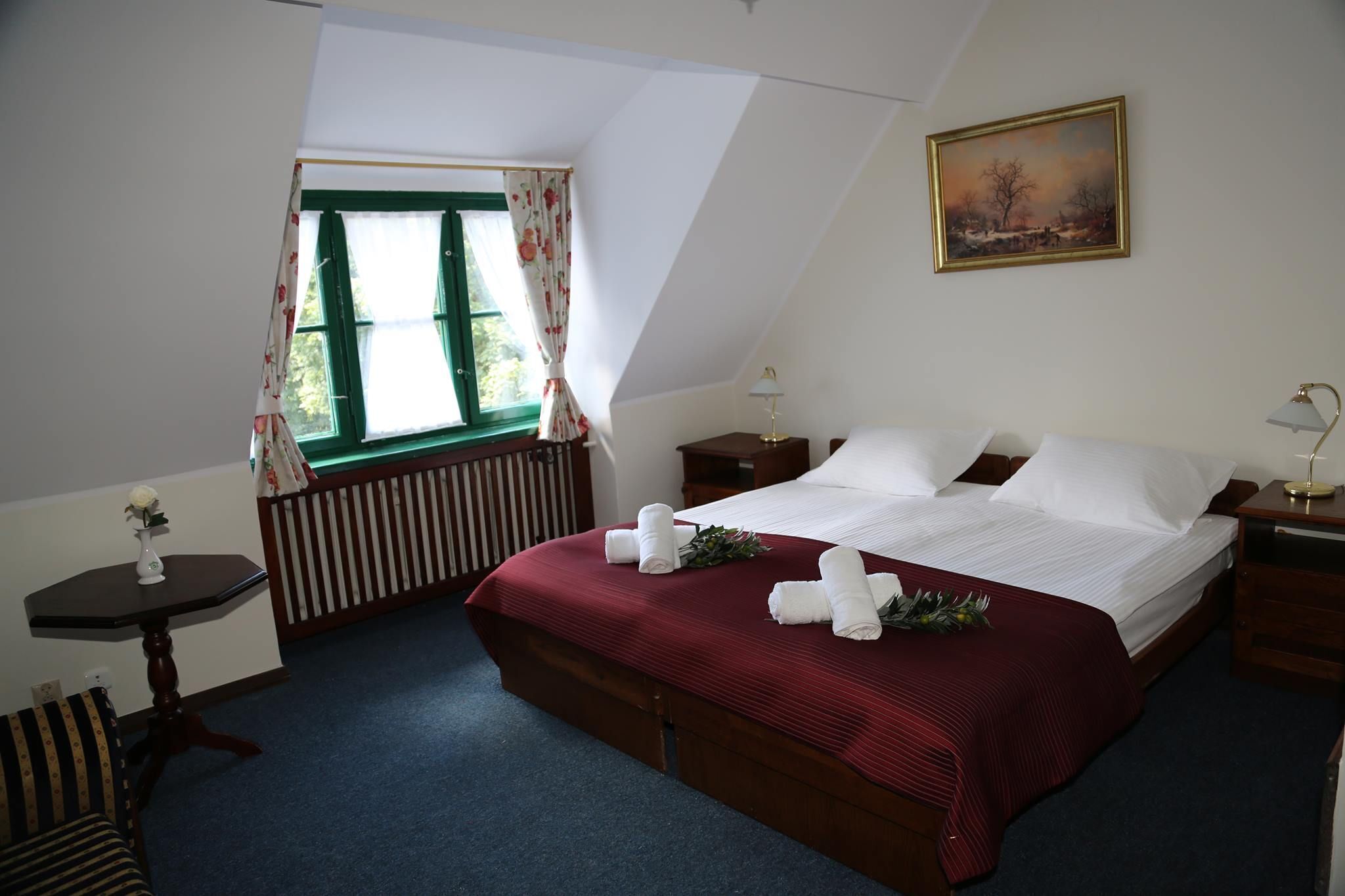 A bedroom with a large bed, a window with curtains, a bedside table with a lamp, and a painting on the wall.