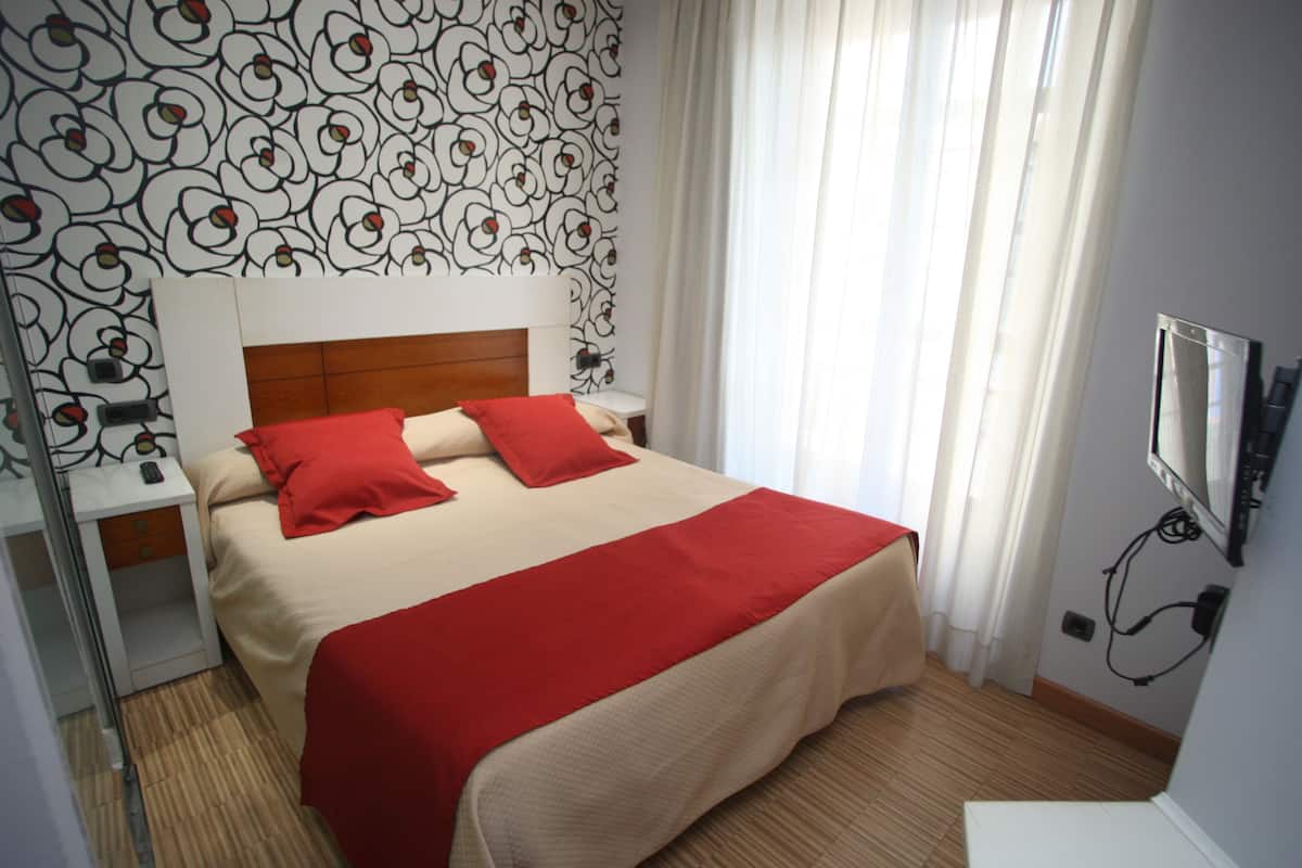 A neatly arranged hotel room with a large bed, red pillows, and a red blanket. There is a wooden floor, a white bedside table, and a wall-mounted TV.