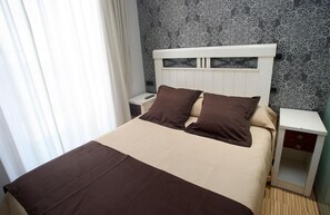 Un lit bien fait, avec une tête de lit blanche, deux oreillers marron et une couverture beige. Il y a une table de chevet blanche avec un tiroir sur le côté droit. La pièce est décorée d’un papier peint à motifs et possède une fenêtre avec des rideaux blancs.