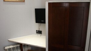 Une petite pièce avec un bureau blanc, un tabouret en bois, un tableau encadré accroché au mur, une porte en bois fermée et un téléviseur noir fixé au mur.