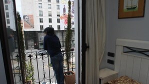 Une chambre d’hôtel avec un balcon, un lit avec une literie à motifs, un tableau encadré au mur et une vue sur un immeuble doté d’une horloge.