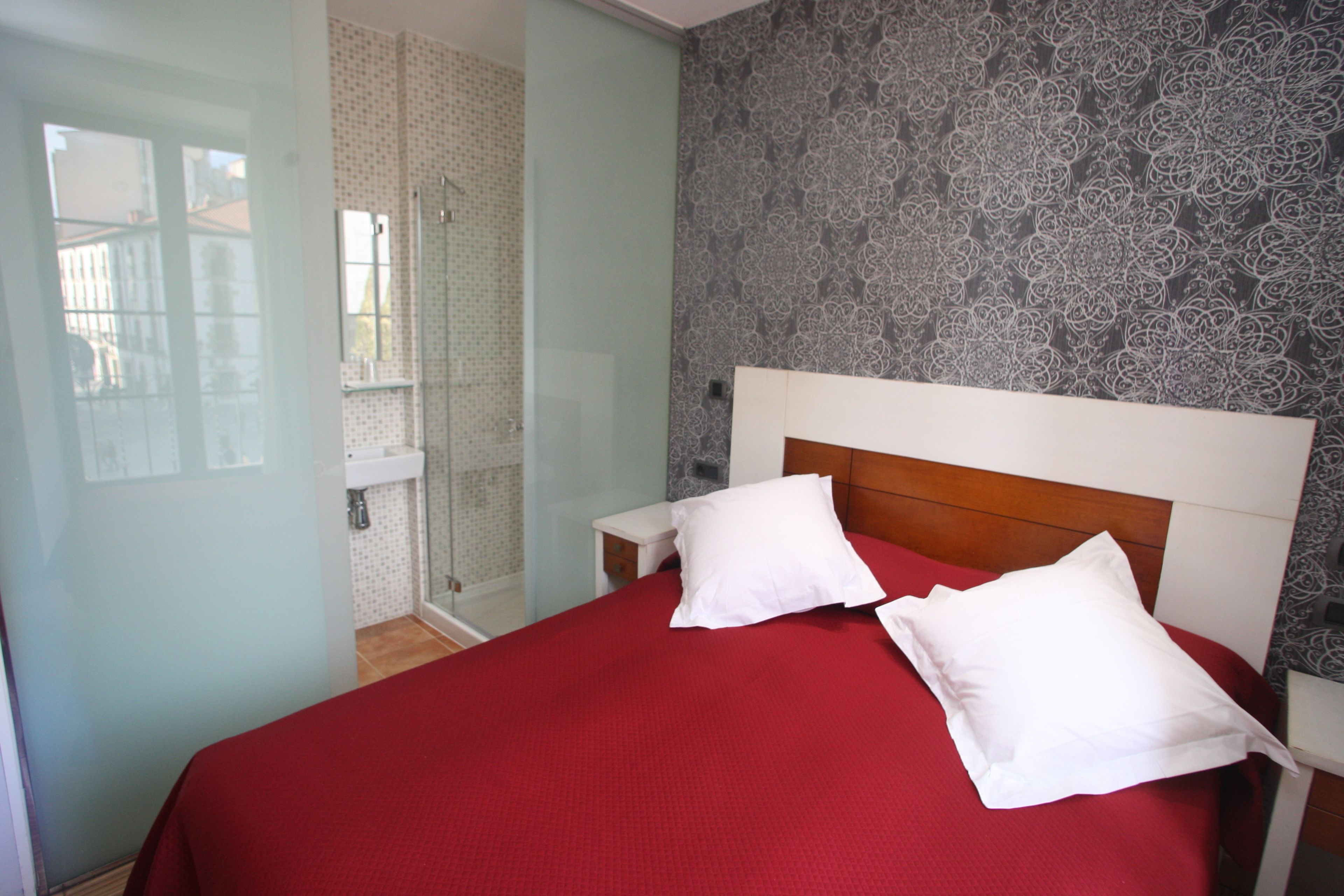 Habitación de hotel con una cama, dos almohadas blancas, cabecera de madera, mesita de noche y baño con ducha y puerta de vidrio.