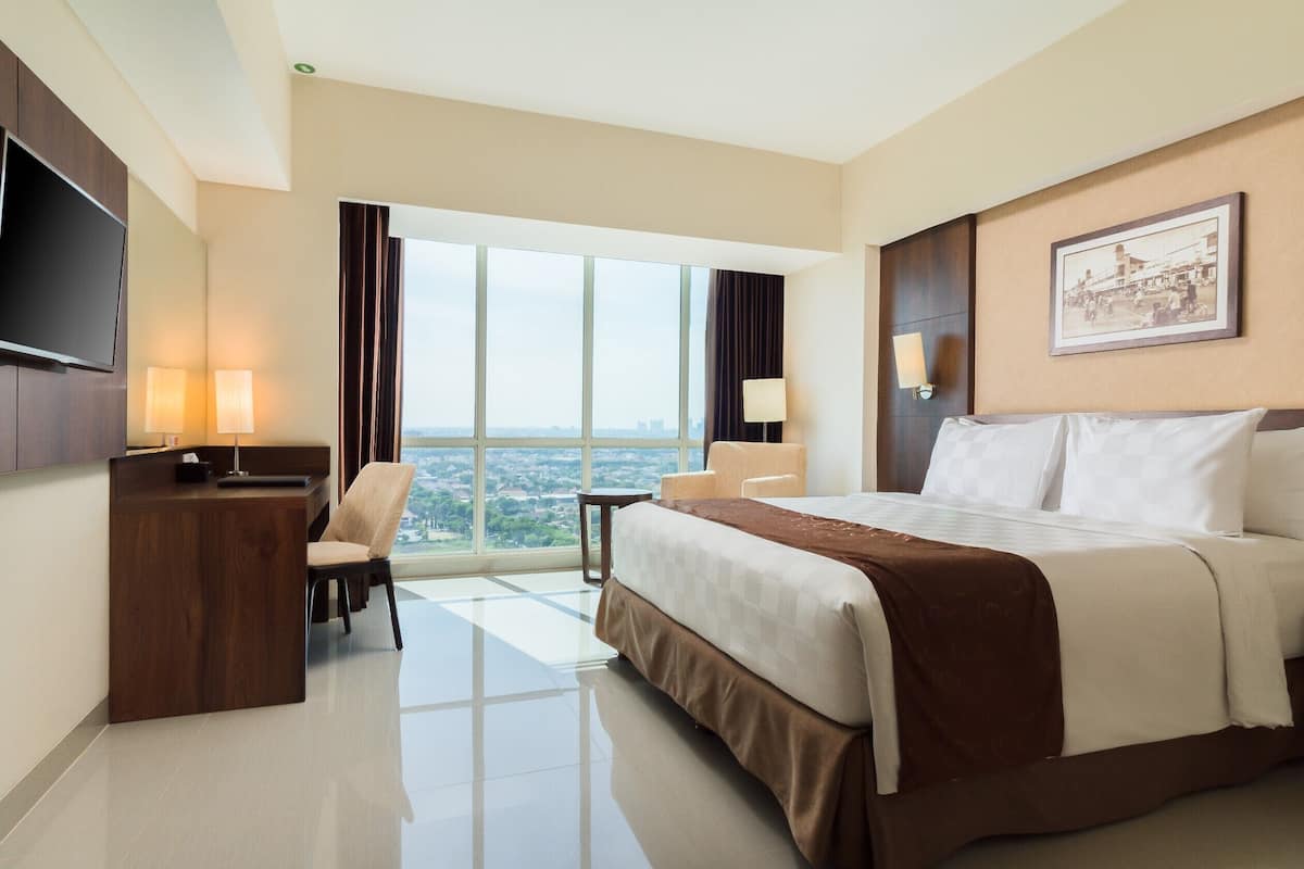 A hotel room with a large bed, a desk with a TV, two chairs, a small table, and a large window offering a city view.