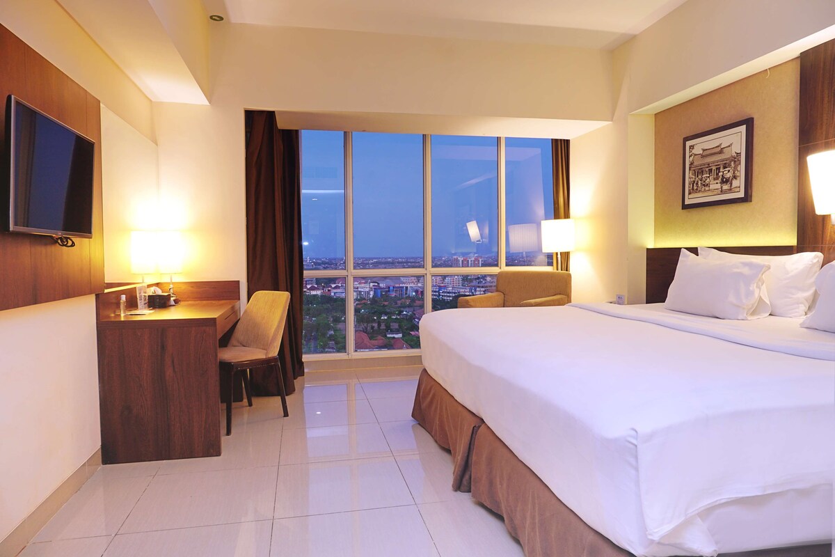Habitación de hotel con una cama grande, un escritorio con silla, televisión y ventana con vista a la ciudad.