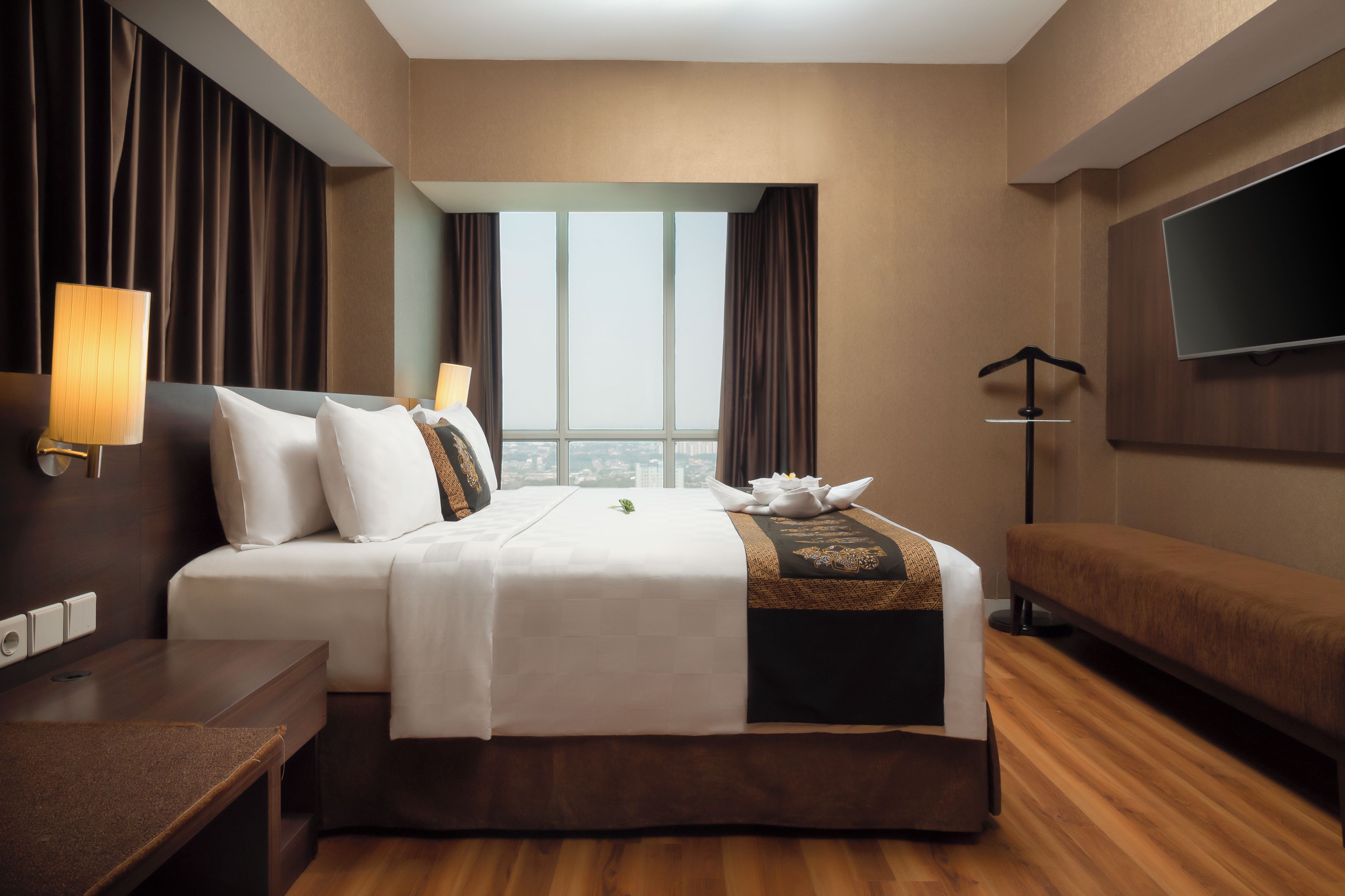 A hotel room with a bed, bedside tables, a desk, a chair, a TV, and a window with a city view.