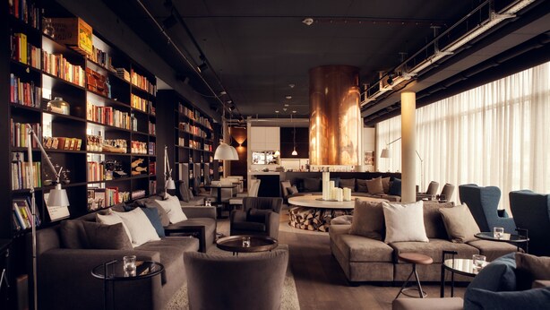 A modern living room with a large bookshelf, comfortable seating, a coffee table, and a large window with curtains. - Soulmade (Garching)