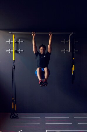 A person performing pull-ups on a bar in a gym with a dark background and exercise equipment. - Soulmade (Garching)
