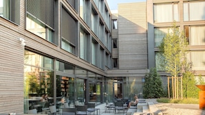 An outdoor seating area with tables and chairs, a pathway, and a building with large windows and a wooden facade. - Soulmade (Garching)