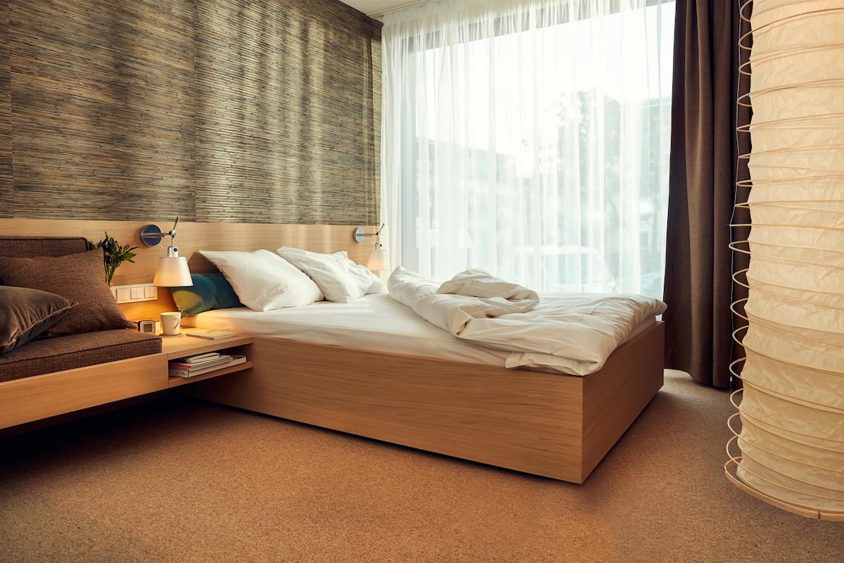 A modern hotel room with a wooden bed, a side table with a lamp, a window with sheer curtains, and a view of buildings outside.