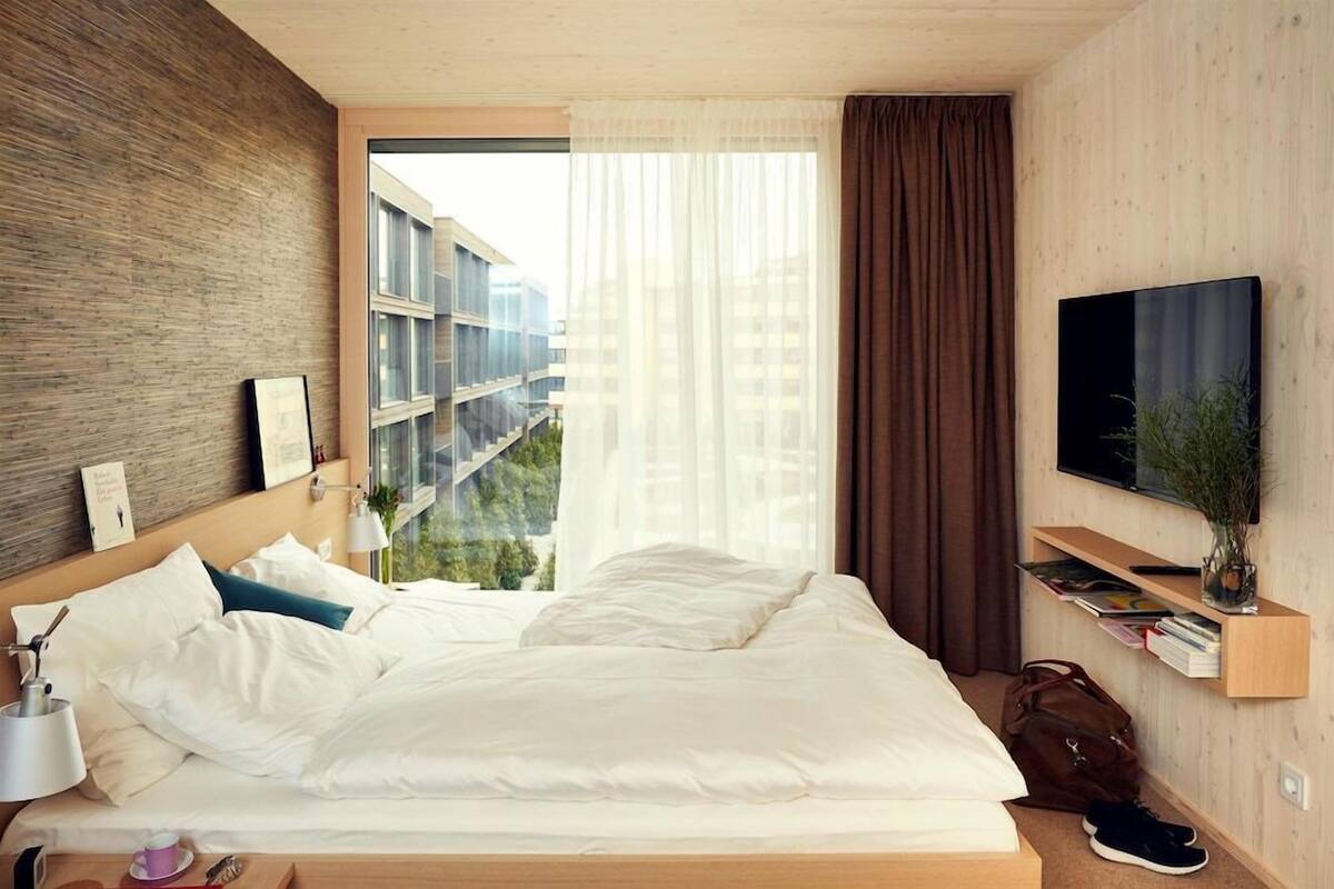 A modern hotel room with a large bed, a TV, a bedside table, a window with curtains, and a view of buildings outside.