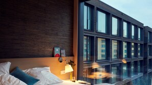 A modern hotel room with a large bed, a bedside table with a lamp, and a view of a building through large windows. - Soulmade (Garching)