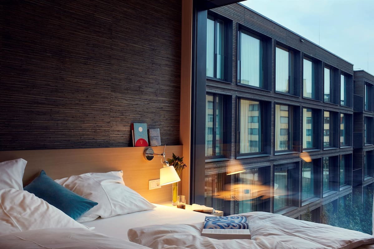 A modern hotel room with a large bed, a bedside table with a lamp, and a view of a building through large windows.