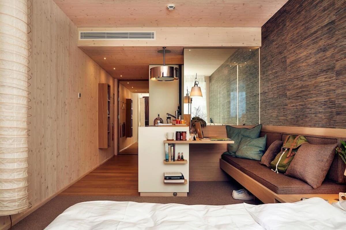 A modern hotel room with a wooden floor, a bed, a sofa, a small desk with a lamp, and a large mirror.