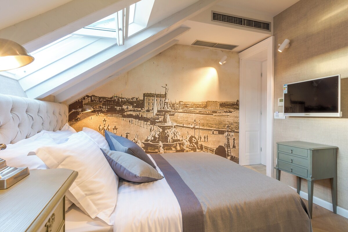 A bedroom with a bed, pillows, a bedside table, a TV, and a wall mural.