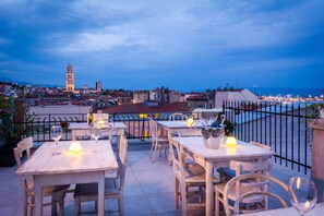 Un espace extérieur aménagé pour les repas, avec des tables et des chaises, offrant une vue sur le paysage urbain, notamment une tour, et une rambarde donnant sur une étendue d’eau.