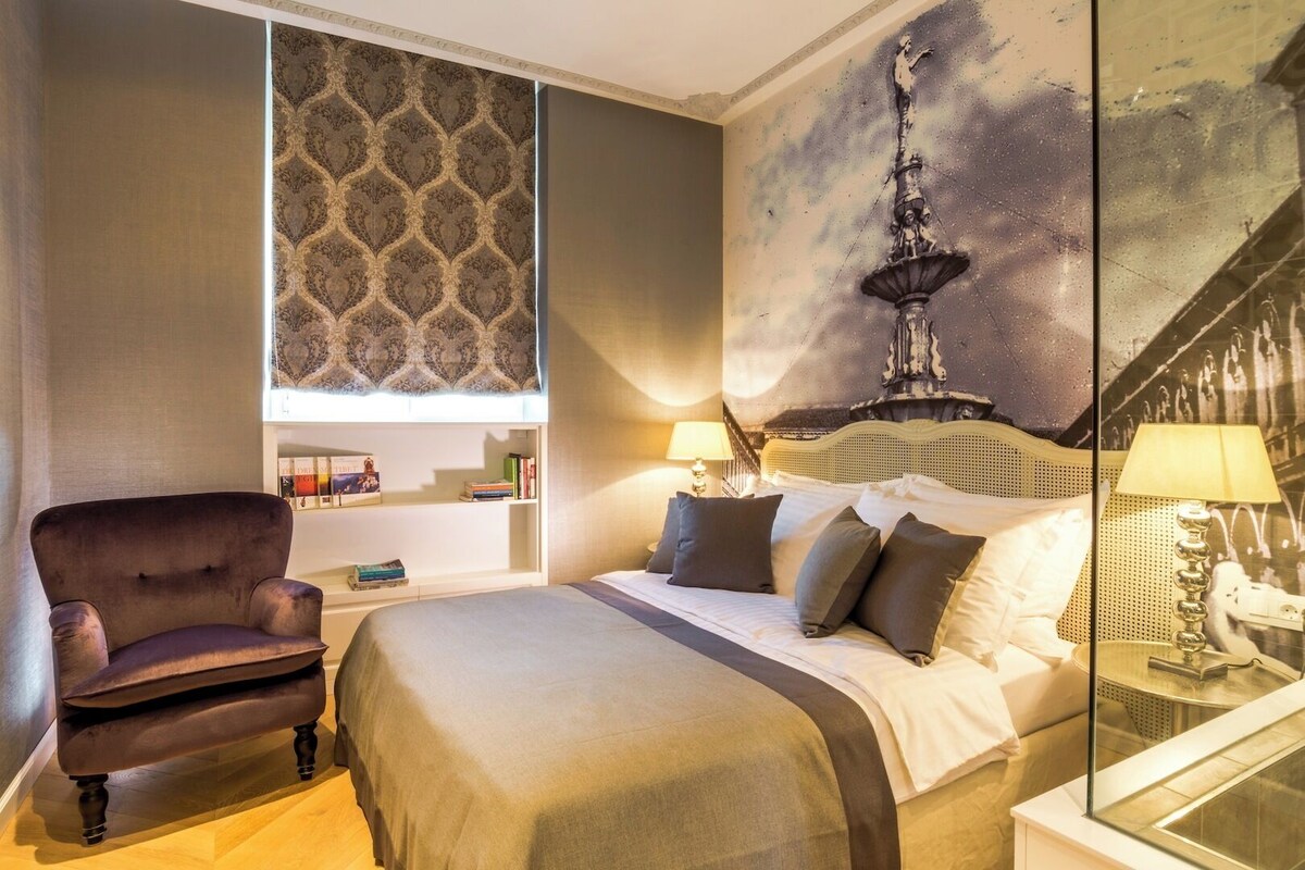 A hotel room with a large bed, a chair, a bookshelf, and a painting of a tower.