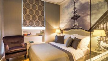 A hotel room with a bed, a chair, a bookshelf, and a large window with a patterned curtain.