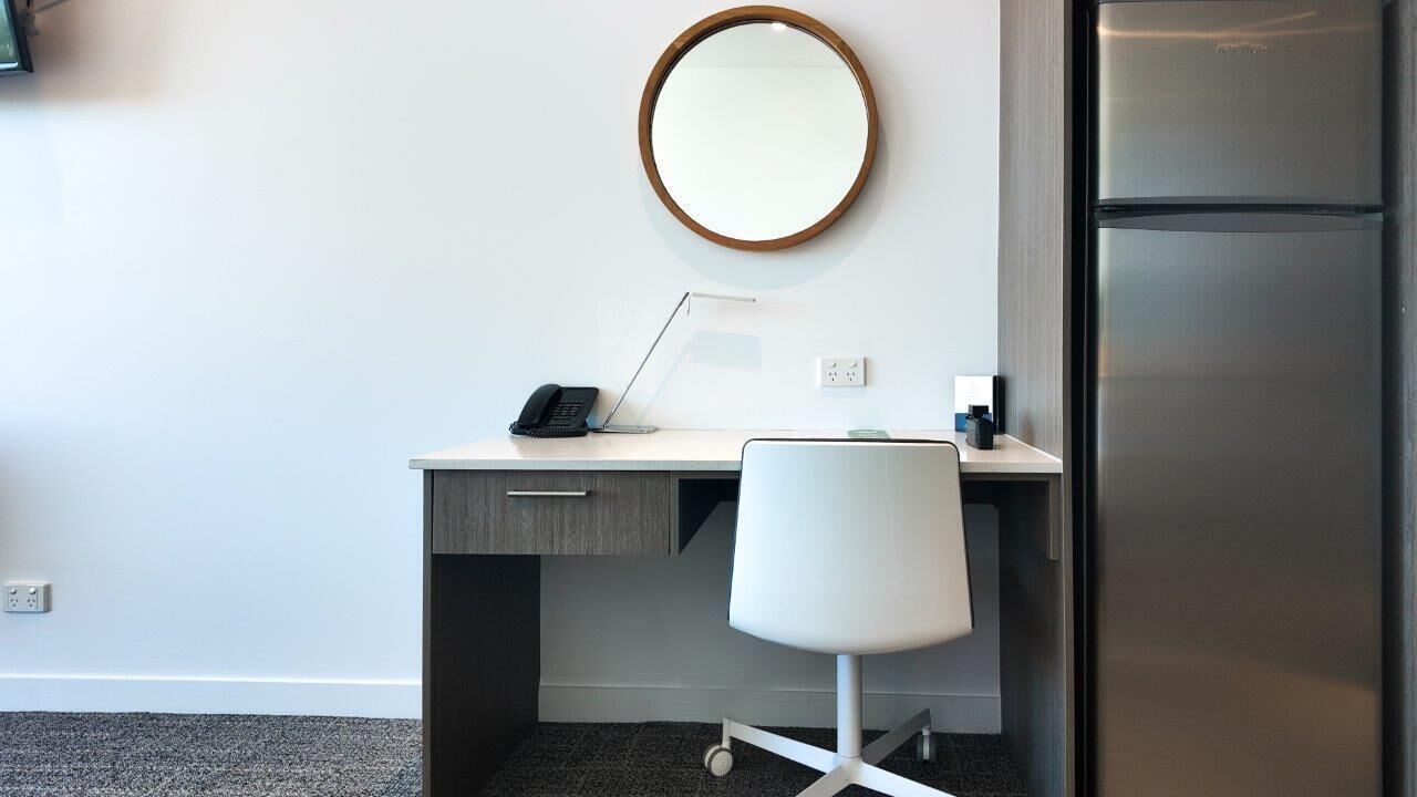 Habitación de hotel con escritorio, silla, espejo, teléfono y refrigerador.