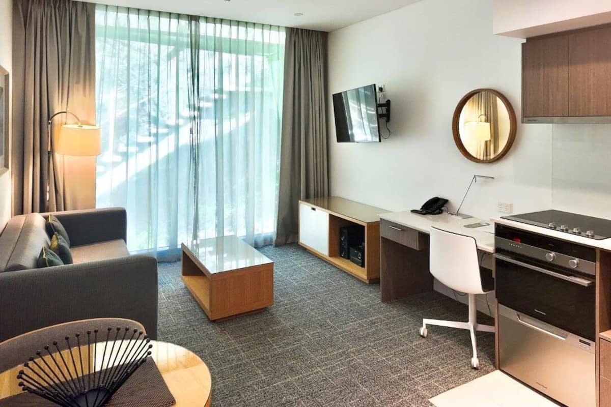 Una habitación de hotel moderna con sofá, escritorio, televisor de pantalla plana, zona de cocina con estufa y horno, y un ventanal con cortinas.