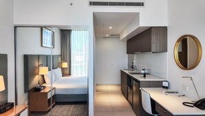 Une chambre d’hôtel comprenant un lit, des lampes de chevet, une table de chevet, un bureau, une chaise, un coin cuisine avec des placards et un évier, ainsi qu’un miroir rond accroché au mur.