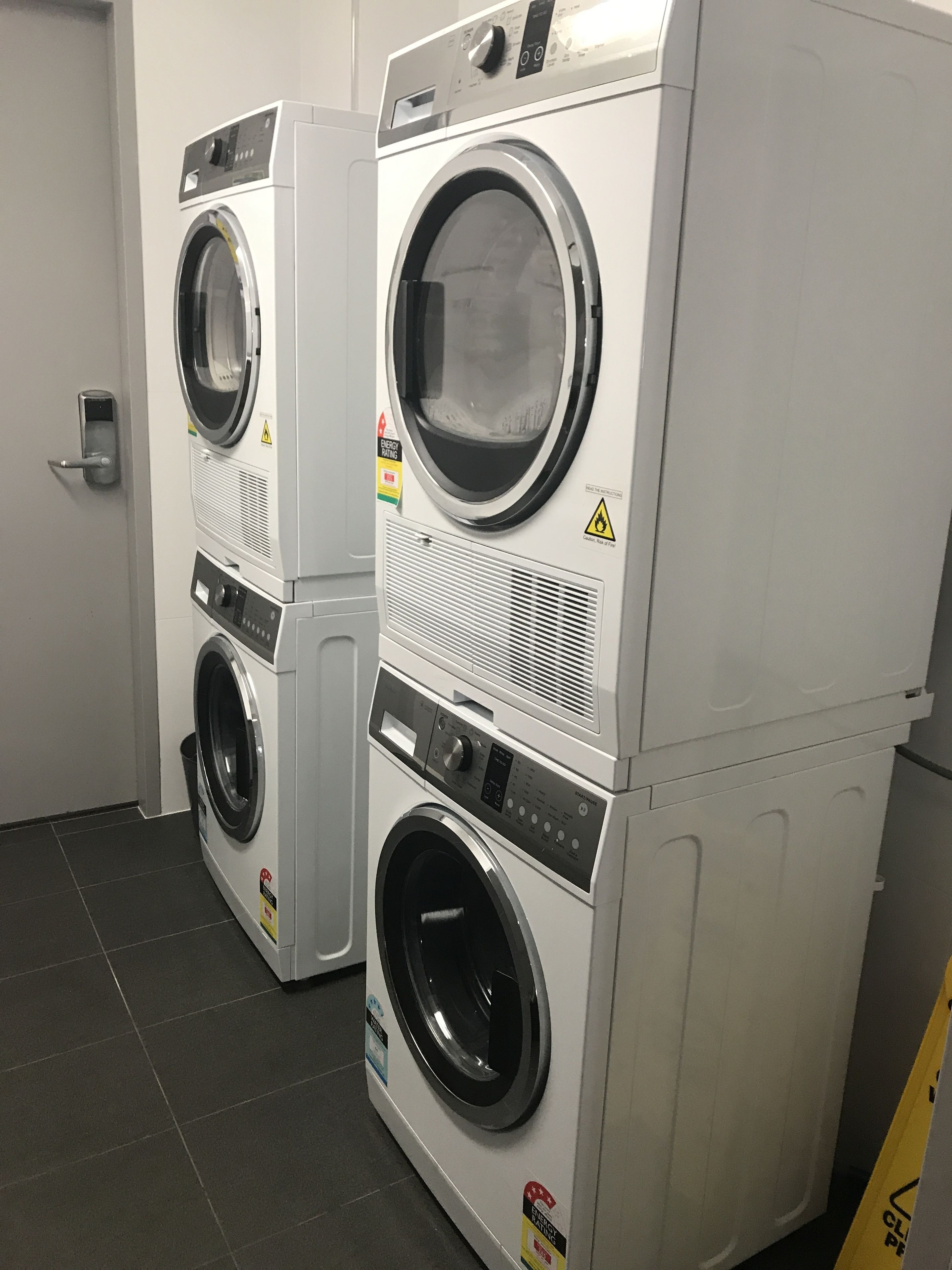 Two stacked front-loading washing machines in a laundry room with a door and a trash bin.