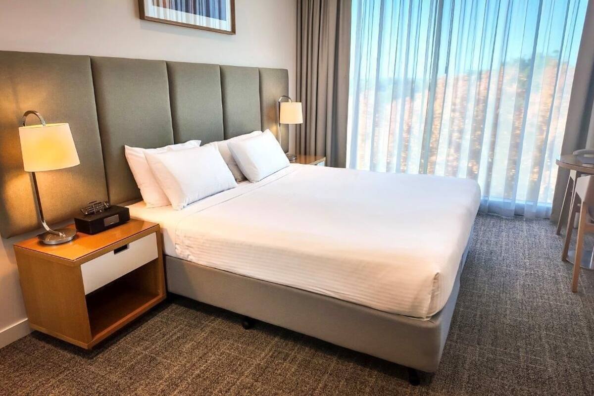 Habitación de hotel con una cama grande, dos lámparas de noche, una mesita de noche con un teléfono y una pequeña mesa, y vistas al exterior a través de cortinas traslúcidas.