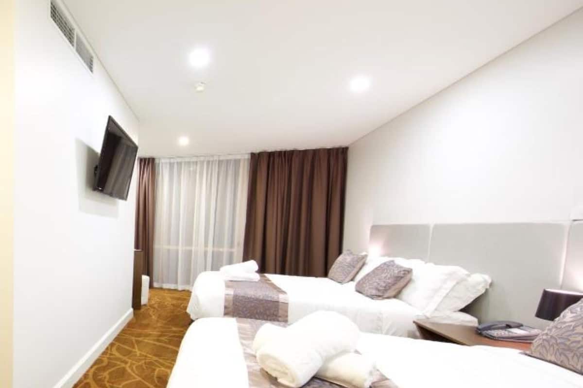 A hotel room with two beds, a flat-screen TV mounted on the wall, a large window with curtains, and a carpeted floor.