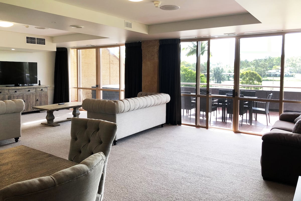 A spacious living room with a sofa, armchairs, a coffee table, and a flat-screen TV. Large sliding glass doors lead to an outdoor area with a dining table and chairs.
