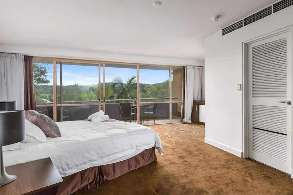 A bedroom with a large bed, a desk, and a balcony with a view of greenery.